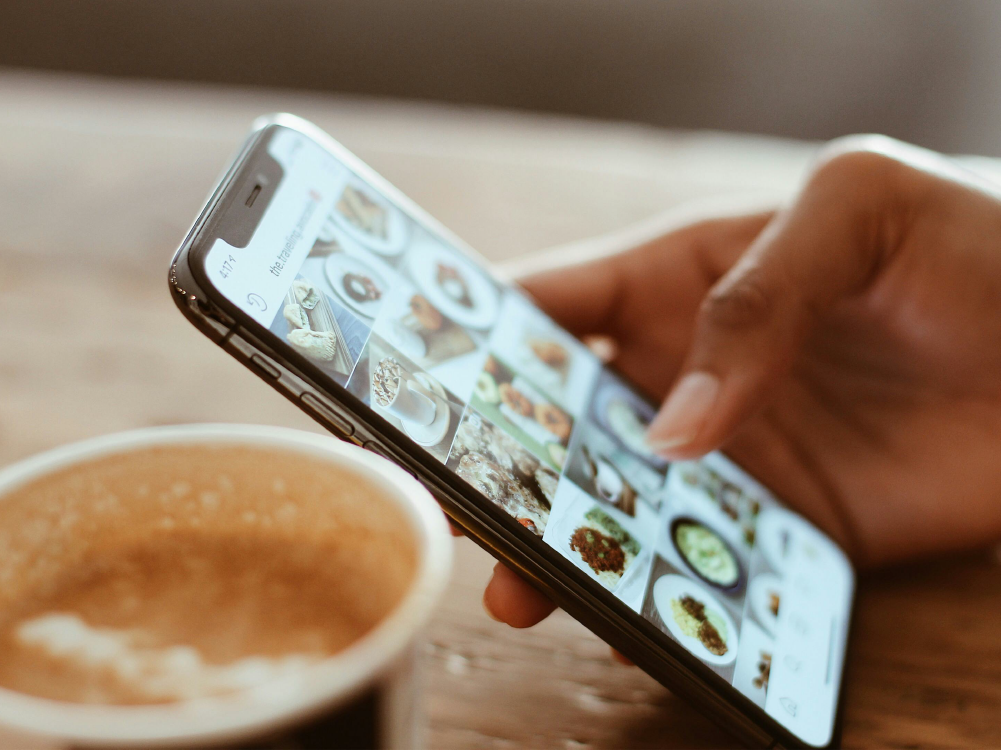 Hand scrolls through food photos on smartphone next to empty coffee cup.